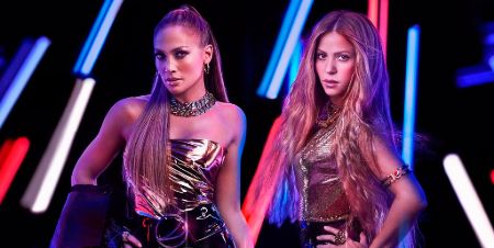 Jennifer Lopez and Shakira posing wearing shiny dresses and blue and red color lights in the background.